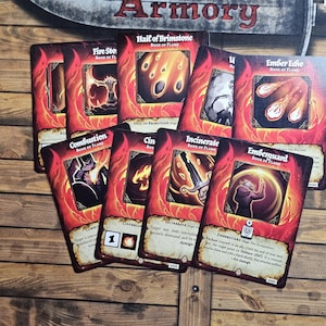 May include: A collection of fantasy-themed game cards with red and orange borders, featuring illustrations of fire and magical elements. The cards have titles like "Fire Stor," "Hail of Brimstone," and "Ember Echo." The cards are arranged on a wooden surface.
