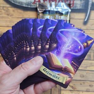 May include: A hand fans a deck of fantasy-themed cards with a purple and gold color scheme. The cards feature illustrations of books, potions, and magical effects. The word "Grimoire" is visible on one card.