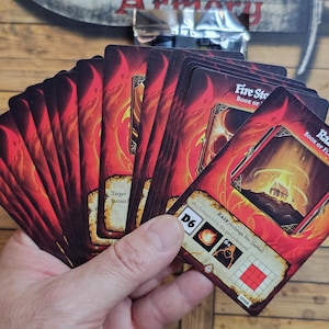 May include: A hand holding a deck of fantasy-themed playing cards. The cards feature fiery red and orange artwork with the text "Fire Stone Book of Flame" and "Raze Book of Flame". The cards have a D6 symbol and other game-related icons.