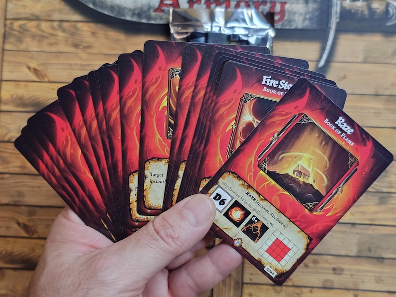 May include: A hand holding a deck of fantasy-themed playing cards. The cards feature fiery red and orange artwork with the text "Fire Stone Book of Flame" and "Raze Book of Flame". The cards have a D6 symbol and other game-related icons.