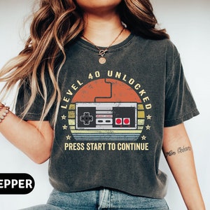 May include: A dark gray t-shirt with a vintage-style graphic of a video game controller. The graphic says "Level 40 Unlocked" and "Press Start to Continue".