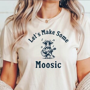 May include: A cream-colored t-shirt with a black and white cartoon cow playing a guitar. The text "Let's Make Some Moosic" is printed on the shirt.