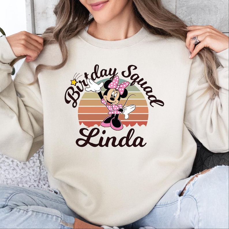 A woman wearing a white sweatshirt with a Minnie Mouse graphic and the text "Birthday Squad Linda" printed on it.