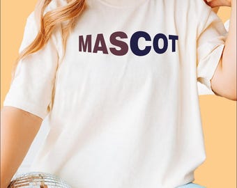 May include: Cream-colored t-shirt with the word "MASCOT" printed across the chest in a gradient of brown and navy blue. The shirt has short sleeves and a relaxed fit, suitable for casual wear.