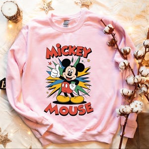 A pink sweatshirt with a Mickey Mouse graphic, along with some cotton balls, a candle, and some star decorations, all arranged on a white surface.