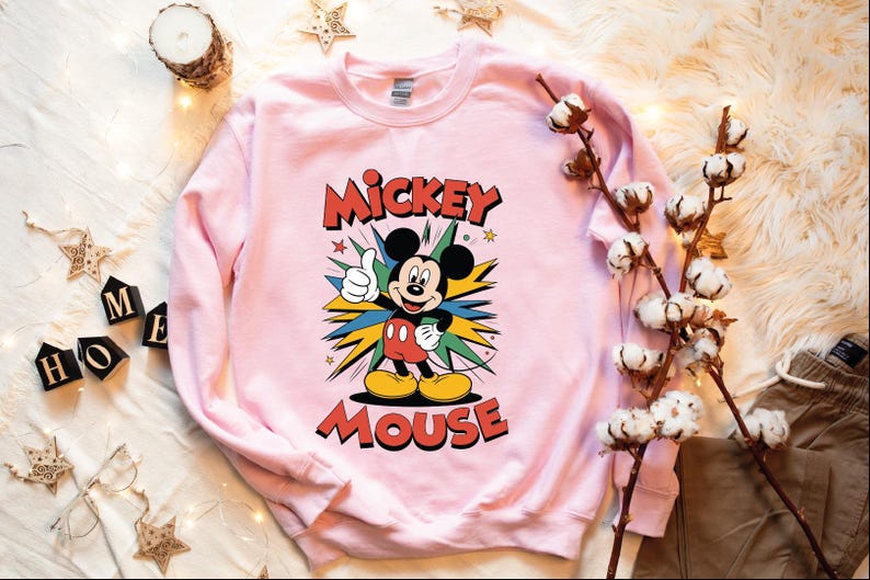 A pink sweatshirt with a Mickey Mouse graphic, along with some cotton balls, a candle, and some star decorations, all arranged on a white surface.