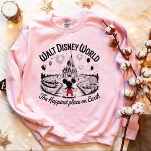 A pink sweatshirt with a Disney World logo and text, along with some cotton balls, a candle, and other decorative items, all arranged on a white surface.