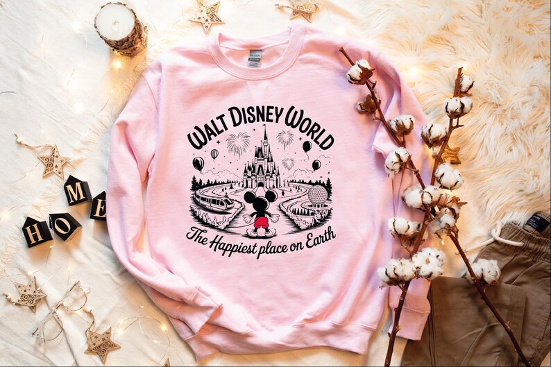 A pink sweatshirt with a Disney World logo and text, along with some cotton balls, a candle, and other decorative items, all arranged on a white surface.