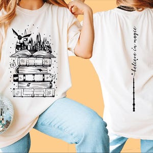 May include: White t-shirt with a black and white illustration of a stack of books, a castle, and a flying owl. The back of the shirt has the text "Believe in magic" with a wand.