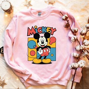 A pink sweatshirt with a Mickey Mouse graphic is displayed on a white surface, surrounded by various objects such as a candle, cotton balls, and star decorations.