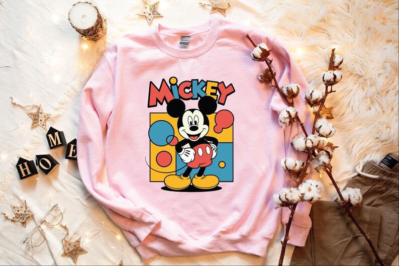 A pink sweatshirt with a Mickey Mouse graphic is displayed on a white surface, surrounded by various objects such as a candle, cotton balls, and star decorations.