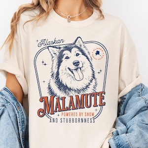 May include: Ivory-colored t-shirt featuring a graphic of an Alaskan Malamute dog. The design includes the word "Alaskan" above the dog, and "Malamute" below, with the phrase "Powered by Snow and Stubbornness".