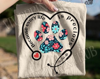 May include: A beige t-shirt featuring a black heart-shaped stethoscope outline surrounding a floral paw print. The text reads Because every life is precious and Vet Tech Life. The paw print is filled with a dark blue background and colorful flowers.