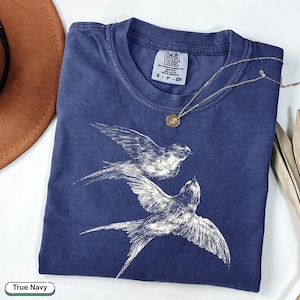 May include: A navy blue short-sleeve t-shirt featuring a white line-art graphic of two flying swallows. The shirt has a crew neckline and a Comfort Colors brand label at the inner collar. A thin gold-tone chain necklace with a small round pendant rests on the fabric.