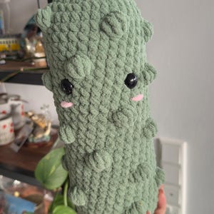Op de afbeelding: Een cilindervormig gehaakt knuffeltje in saliegroen, met een bobbelige textuur, twee ronde zwarte plastic ogen en kleine roze geborduurde wangen. Het zachte, handgemaakte item heeft een afgeronde boven- en onderkant en lijkt op een augurk.
