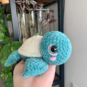 Può includere: Una tartaruga di peluche fatta a mano nei toni del blu e del crema. La tartaruga ha grandi occhi glitterati, una bocca rosa e un aspetto morbido e strutturato. La tartaruga è un giocattolo carino e coccoloso.