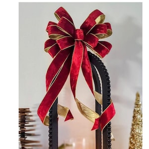 May include: A black metal lantern with a red velvet bow and gold ribbon on top. The lantern has a glass insert with a lit candle inside. The lantern is surrounded by greenery.