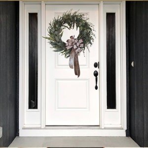 May include: A white front door with a green and brown eucalyptus wreath with a brown velvet bow. The wreath is hanging on the door with a black door handle.