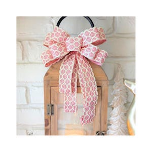 May include: A wooden lantern with a large pink bow. The bow has a white geometric pattern and long, flowing tails. The lantern has a black handle and a glass front, perfect for home decor.