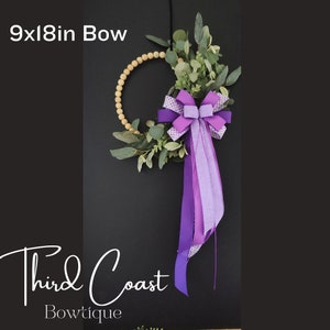 May include: A 9x18 inch wooden bead wreath with a purple and white checkered bow. The bow has a purple ribbon with white polka dots hanging from it. The wreath is decorated with green eucalyptus leaves.