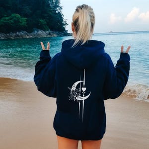 Può includere: Felpa con cappuccio blu navy con un design bianco a mezzaluna, floreale e a cuore sul retro. La felpa ha un cappuccio con coulisse e maniche lunghe. La persona è su una spiaggia sabbiosa con l'oceano sullo sfondo.