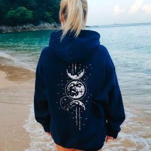 Könnte beinhalten: Ein marineblauer Hoodie mit einem weißen Mondphasen-Design auf der Rückseite. Das Design zeigt drei Monde, Sterne und eine vertikale Linie mit geometrischen Formen.