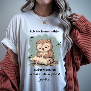 Könnte beinhalten: Weißes T-Shirt mit einem Cartoon-Eulen-Design, die auf einem Kissen liegt, mit dem Text "Ich bin immer müde, außer wenn ich schlafe... dann gehts!" und der Signatur "Jeandra". Die Eule ist braun und beige, vor hellgrünem Hintergrund.