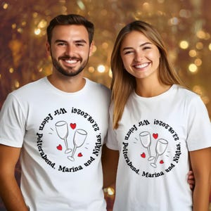 Könnte beinhalten: Zwei Personen tragen weiße T-Shirts mit einem kreisförmigen Design. Das Design umfasst zwei Weingläser, rote Herzen und Text auf Deutsch: "Wir feiern Polterabend, Marina und Niklas, 23.08.2026!"