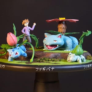 Puede incluir: Figura detallada con múltiples personajes de Pokémon y un entrenador. La pieza central es un gran Venusaur azul con la boca abierta, rodeado de figuras más pequeñas de Bulbasaur y un entrenador. La base tiene texto japonés.