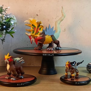 Peut inclure: Ensemble de trois figurines Pokémon détaillées. La figurine centrale est une grande créature multicolore avec une crinière flamboyante, sur une plateforme ronde. Deux figurines plus petites l'entourent, chacune sur des plateformes individuelles. Du texte japonais est visible.