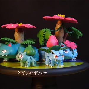 Könnte beinhalten: Detailliertes Figurenset mit mehreren Bulbasaur- und Venusaur-Figuren mit rosa und gelben blütenartigen Strukturen. Die Figuren befinden sich in verschiedenen Posen auf einer runden Basis mit japanischem Text.
