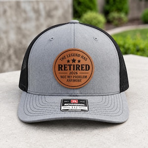 May include: A grey and black trucker hat featuring a circular tan leather patch on the front. The patch is engraved with three stars and the text: THE LEGEND HAS RETIRED 2026 NOT MY PROBLEM ANYMORE. A black rectangular sticker is on the brim.