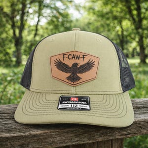 May include: Olive green and black trucker hat with a leather patch featuring a raven and the text "F-CAW-F". The hat has a curved brim and adjustable strap. The Richardson brand label is visible.