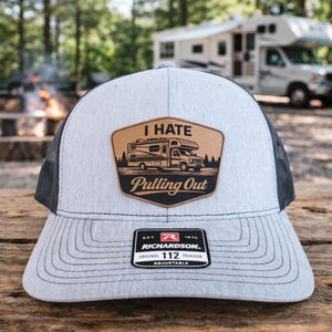 May include: Gray and black trucker hat with a patch that reads "I HATE Pulling Out" with a graphic of a camper. The hat has a curved brim and a mesh back. The Richardson brand label is visible.