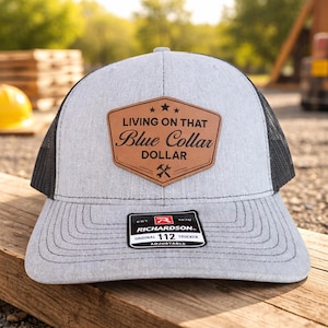 May include: A gray trucker hat with a black mesh back and a curved brim. The hat features a brown leather patch with the text "LIVING ON THAT Blue Collar DOLLAR" and a crossed wrench graphic. The Richardson brand label is visible.