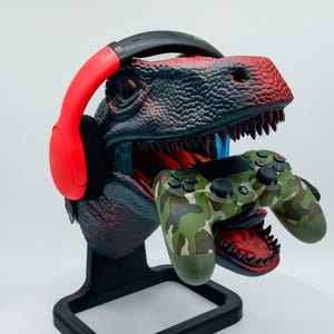 Könnte beinhalten: Ein schwarzer und roter Dinosaurierkopf-Ständer mit einem roten Headset und einem Tarnmuster-grünen Videospiel-Controller. Der Dinosaurier hat den Mund offen und das Headset und der Controller liegen im Maul.