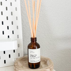 May include: A brown glass reed diffuser bottle with a white label that reads "White Tea & Thyme." The bottle is filled with a clear liquid and topped with several light-colored reeds. The diffuser sits in a wooden bowl on a white surface.