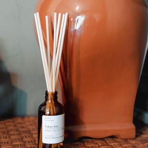 May include: A brown glass reed diffuser bottle with a white label reading Tokyo Zen, containing several light-colored wooden reeds extending upwards. The bottle is cylindrical with a narrow neck and a flat base, displaying clear black text on the label.