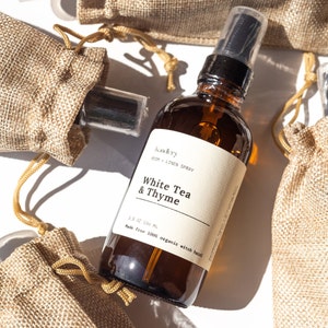 May include: A brown glass bottle with a black spray nozzle filled with a clear liquid. The bottle has a white label with the text "Akry ROOM + LINEN SPRAY White Tea & Thyme 3.5 OZ 100 ML Made from 100% organic witch hazel".