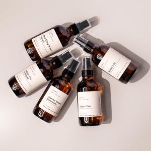 May include: Five amber glass spray bottles with black spray nozzles and white labels. The labels read "Sparkling Water Neroli", "White Tea & Thyme", "Matcha Honey Lemonade", "Santal Life", and "Tokyo Zen". The bottles are arranged on a light gray surface.