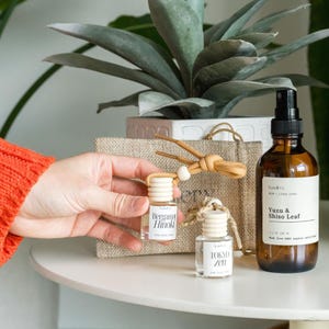 May include: A collection of aromatherapy products, including two small glass bottles labeled "Bergamot Hinoki" and "Tokyo Zen", and a larger amber glass bottle of "Yuzu & Shiso Leaf" room and linen spray. The products are displayed with a decorative plant.