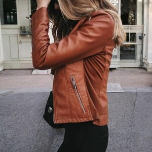 May include: A brown leather jacket with a zip fastening and a zipped pocket on the left side. The jacket is being worn by a person with long blonde hair.