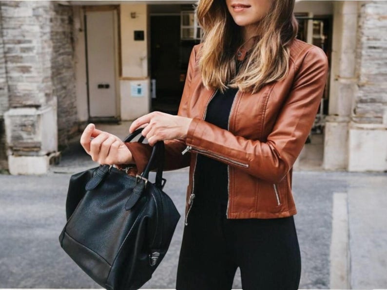 May include: A woman wearing a brown leather jacket and black trousers is holding a black leather handbag.