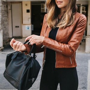 May include: A woman wearing a brown leather jacket and black trousers is holding a black leather handbag.