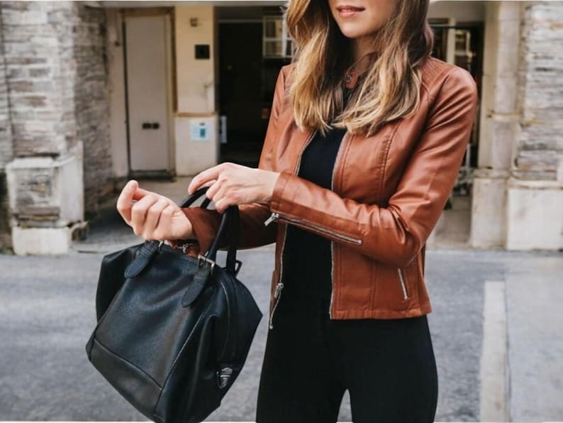 May include: A woman wearing a brown leather jacket and black trousers is holding a black leather handbag.