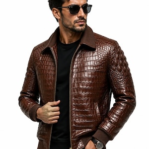 Könnte beinhalten: Eine braune Lederjacke mit texturiertem Krokodilleder-Muster, durchgehendem Frontreißverschluss, geripptem Strickkragen und gerippten Strickbündchen. Die Jacke wird über einem schlichten schwarzen Shirt getragen, kombiniert mit einer schwarzen Hose.