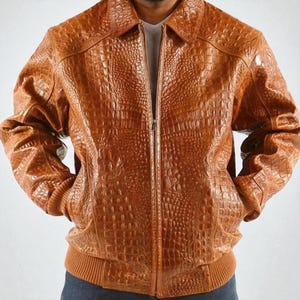 Puede incluir: Chaqueta de cuero marrón con textura de piel de cocodrilo. La chaqueta tiene cuello, cierre de cremallera y puños y dobladillo de canalé. La chaqueta es de color marrón medio.
