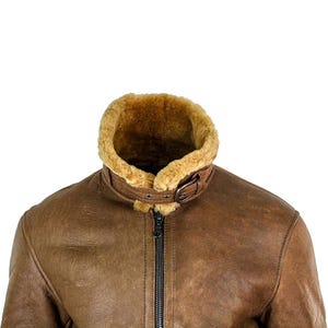 May include: Brown leather jacket with a shearling collar and lining. The jacket features a buckled collar strap and a black zipper closure. The leather has a distressed appearance, and the shearling is a light brown color.