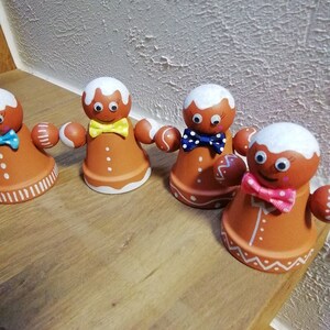 Könnte beinhalten: Vier Lebkuchenmann-Figuren aus Terrakotta-Töpfen, verziert mit weißem Zuckerguss, Glitzern und bunten Schleifen. Die Figuren sind auf einer Holzoberfläche angeordnet.