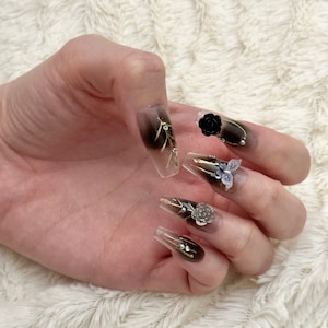 May include: Close-up of a hand wearing long, clear, brown ombre press-on nails with gold accents and black and silver 3D floral embellishments.